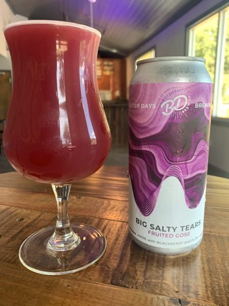 A Big Salty Tears fruited gose can next to a tulip glass of the deep red beer on a wooden table