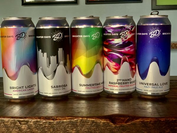 Five Brighter Days cans lined up on a wooden bar: Bright Lights, Sabrosa, Summertime Magic, 2nd Hand Raspberry Beret and Universal Love